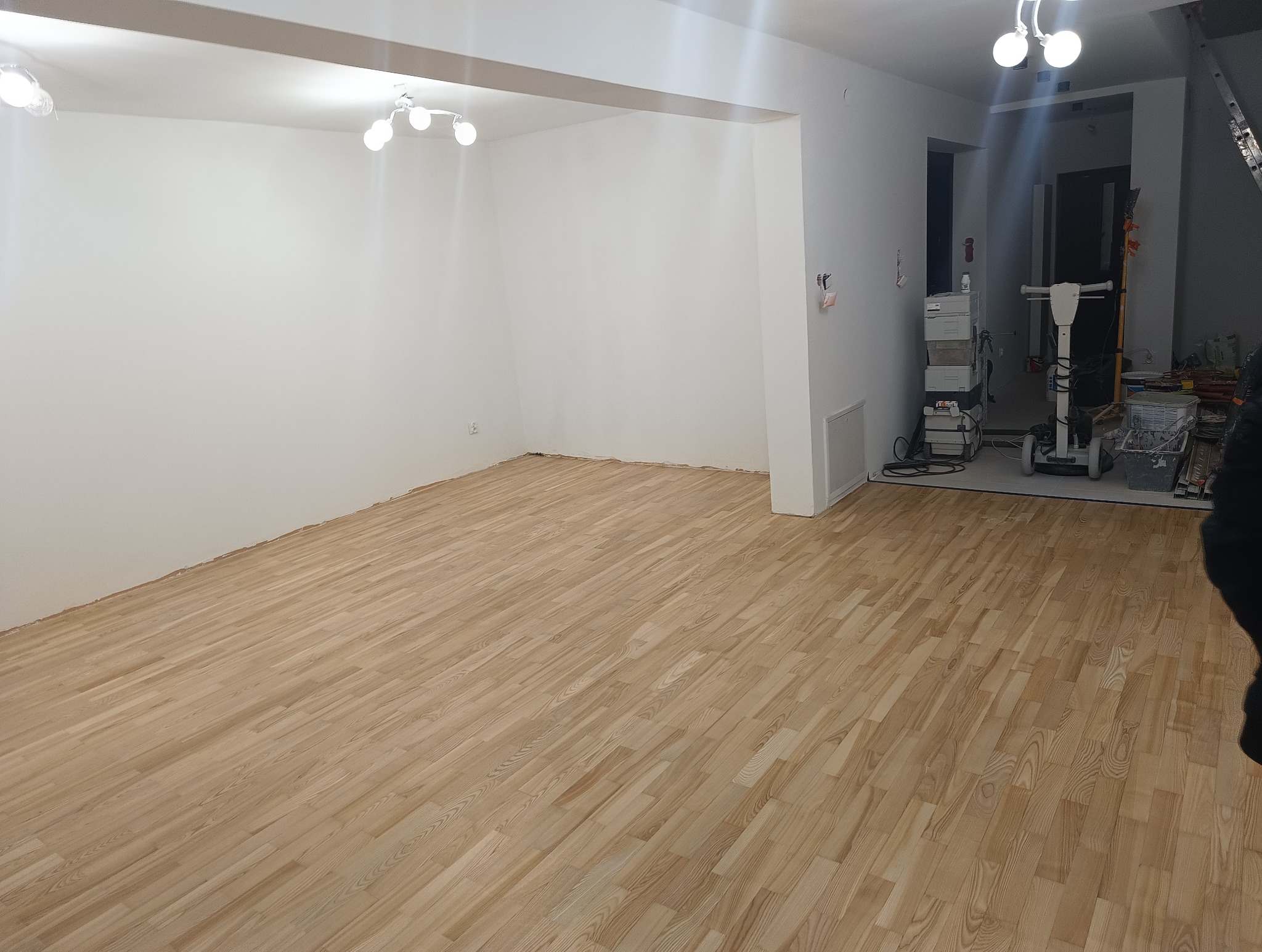 Jasna, odnowiona podłoga parkietowa w pustym pomieszczeniu z białymi ścianami. W tle widoczna maszyna do obróbki drewna i narzędzia.