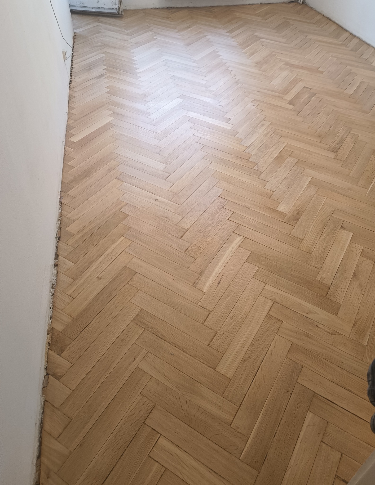 Parkiet ułożony w jodełkę po renowacji. Jasne drewno, widoczne detale słojów. Świeżo wykończona podłoga w pomieszczeniu z białymi ścianami.