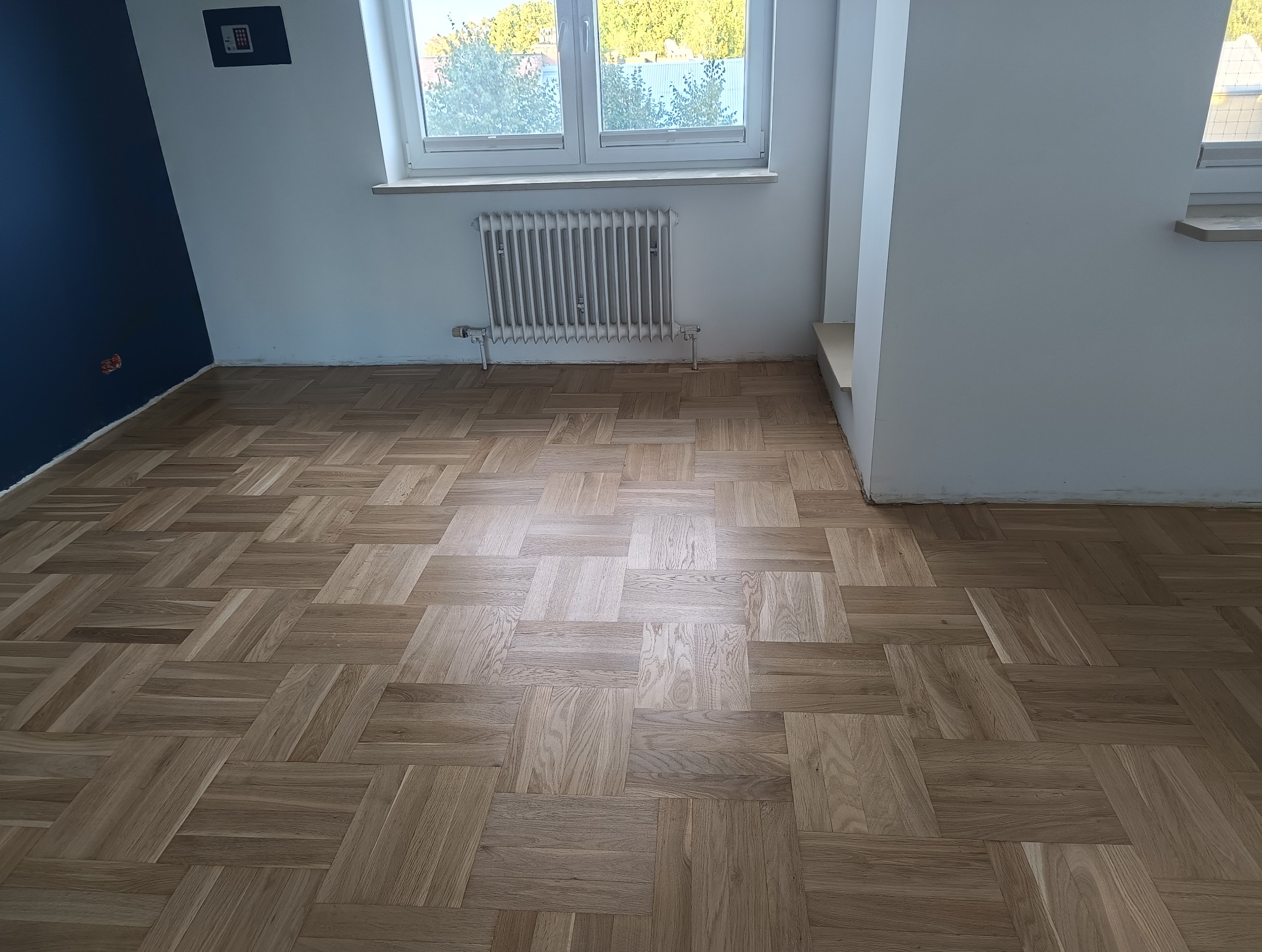 Parkiet dębowy ułożony w jodełkę francuską, po renowacji. Widoczne naturalne usłojenie drewna i świeży lakier. W tle białe ściany i grzejnik.