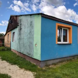 Niebieski budynek z pomarańczowymi obramowaniami okien, widziany z perspektywy ogrodu w słoneczny dzień. Widoczna antena satelitarna na dachu.