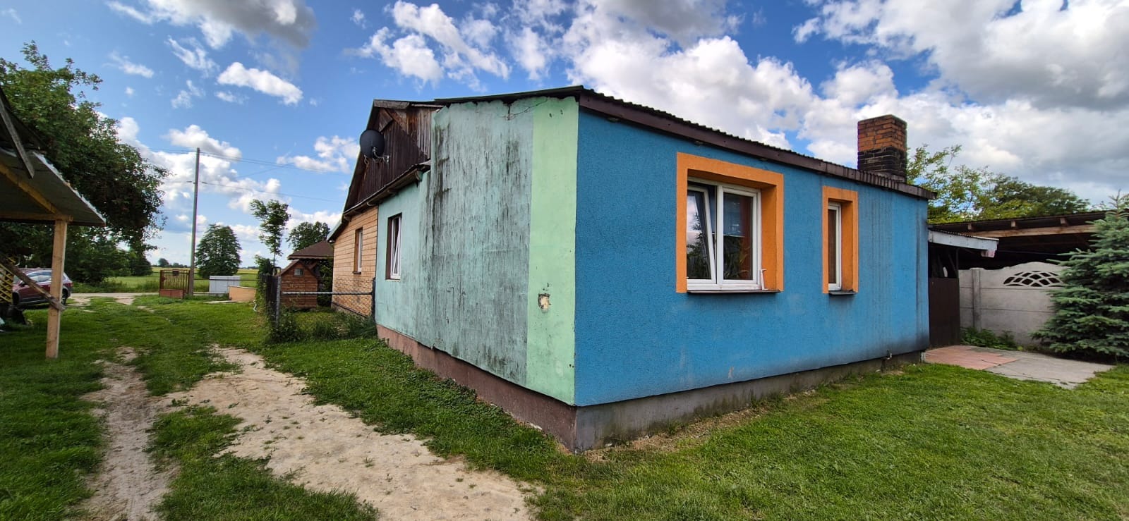 Niebieski budynek z pomarańczowymi obramowaniami okien, widziany z perspektywy ogrodu w słoneczny dzień. Widoczna antena satelitarna na dachu.