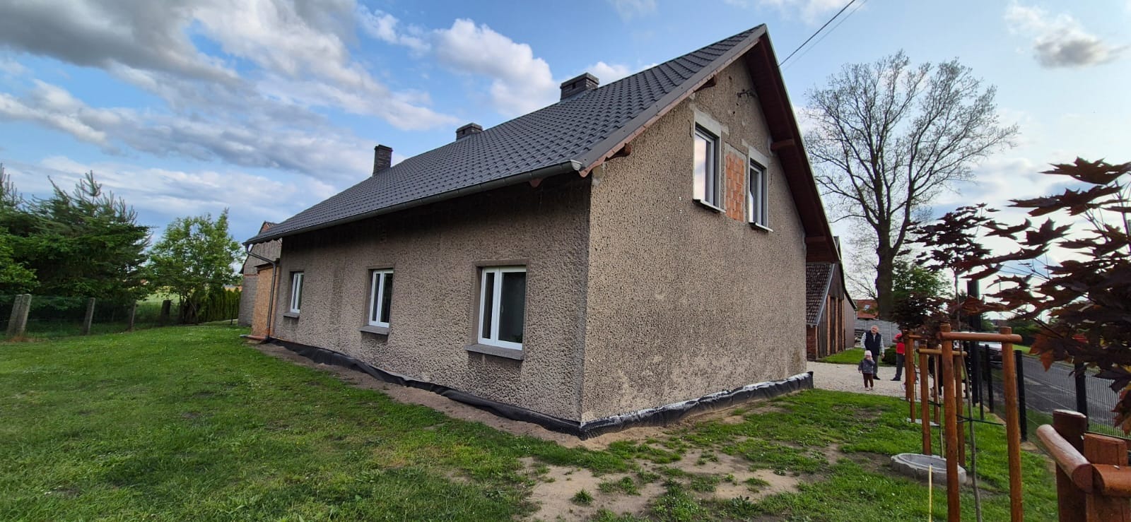 Dom jednorodzinny z szarą elewacją i ciemnym dachem, widok z boku na tle zielonego trawnika i błękitnego nieba z chmurami. Częściowo wymienione okna.