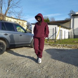 Val.Dav Fashion - Modelka prezentuje bordowy, dresowy komplet z kapturem na zewnątrz, obok zaparkowanego srebrnego SUV-a, w słoneczny dzień. Widoczne budynki w tle.