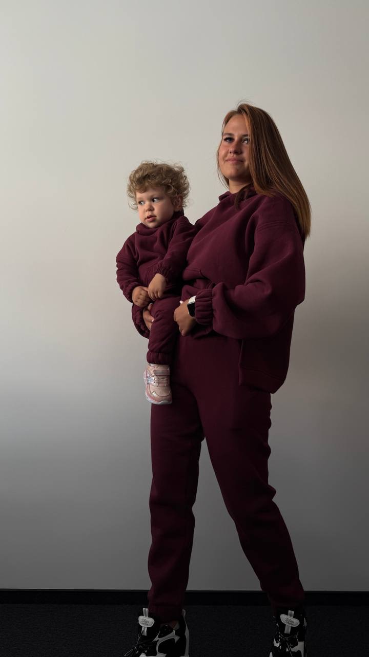 Matka trzymająca dziecko, oboje w bordowych, dresowych kompletach. Mama ma długie, proste włosy, dziecko kręcone. Widoczne buty mamy, czarno-białe sneakersy.