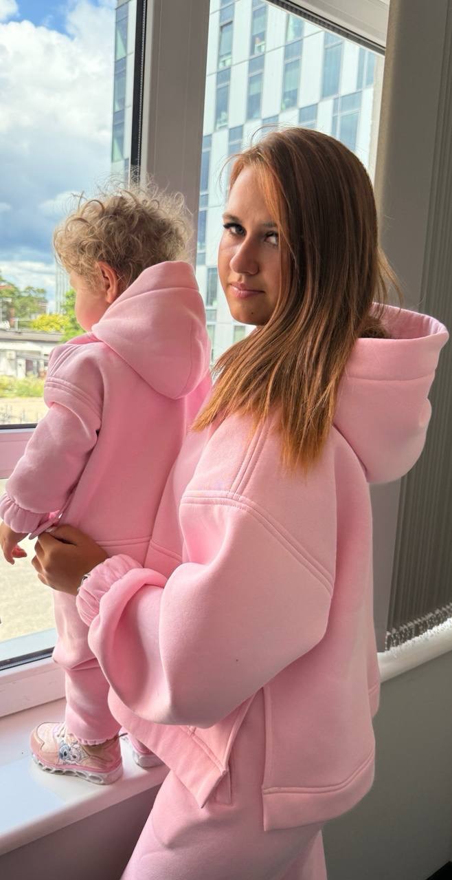Mama z dzieckiem w identycznych, różowych dresach z kapturem, stoją przy oknie z widokiem na miasto. Dresy mają charakterystyczne rozcięcia na bokach bluzy.