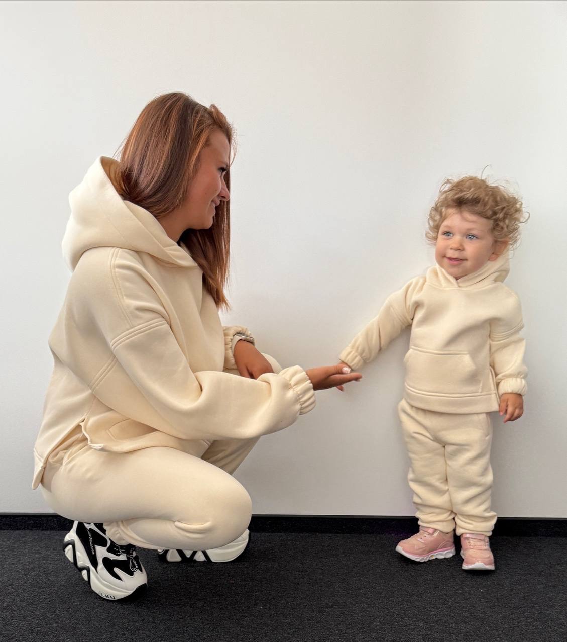 Mama i syn w beżowych dresach z kapturem, trzymają się za ręce na tle białej ściany. Ona kuca, on stoi. Uwagę przyciągają masywne buty mamy i kręcone włosy synka.
