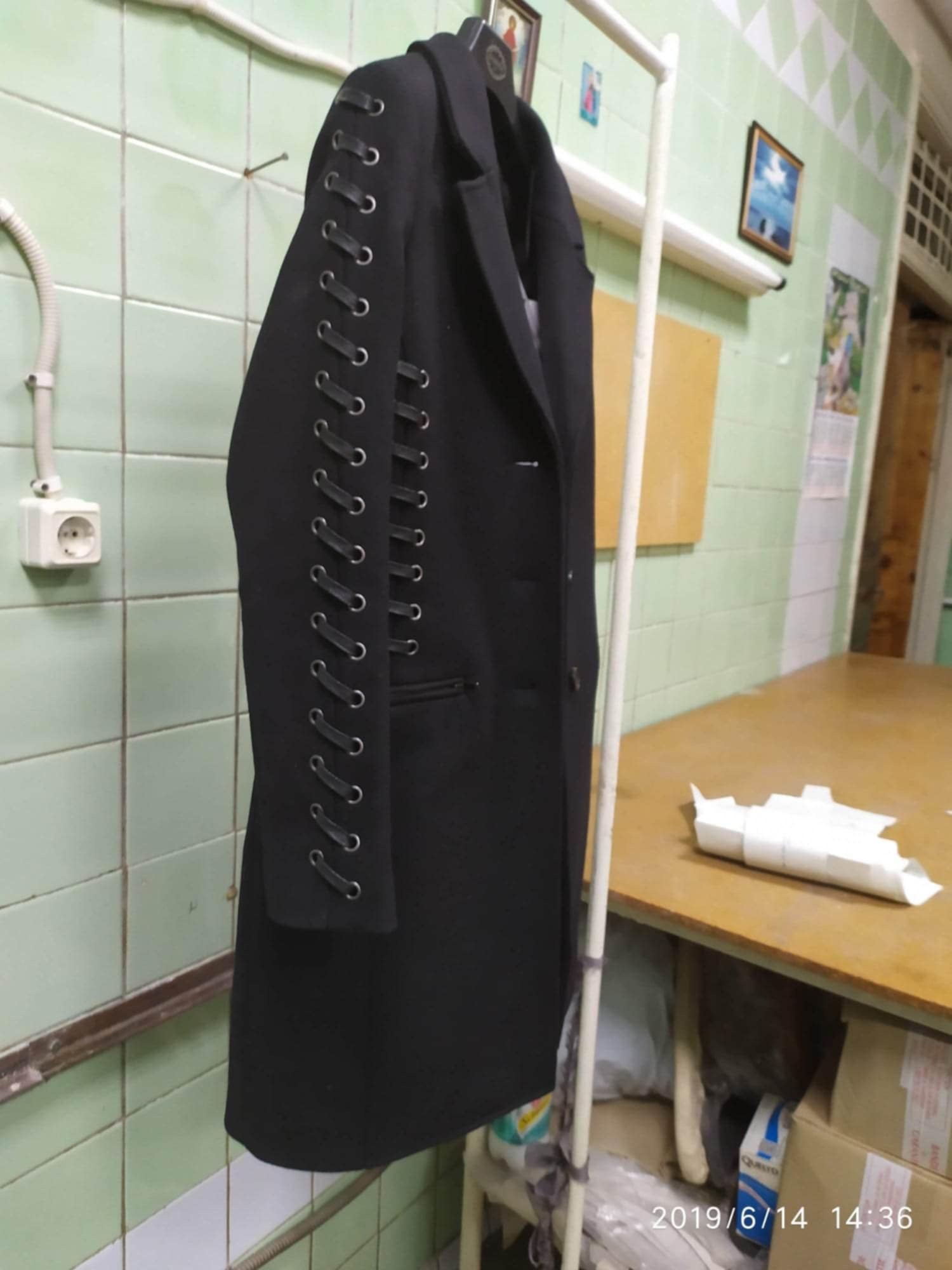 Ciemny, damski płaszcz z ozdobnymi szlufkami na rękawach, wiszący na wieszaku w szwalni. Widoczne płytki ceramiczne na ścianie i stół roboczy.