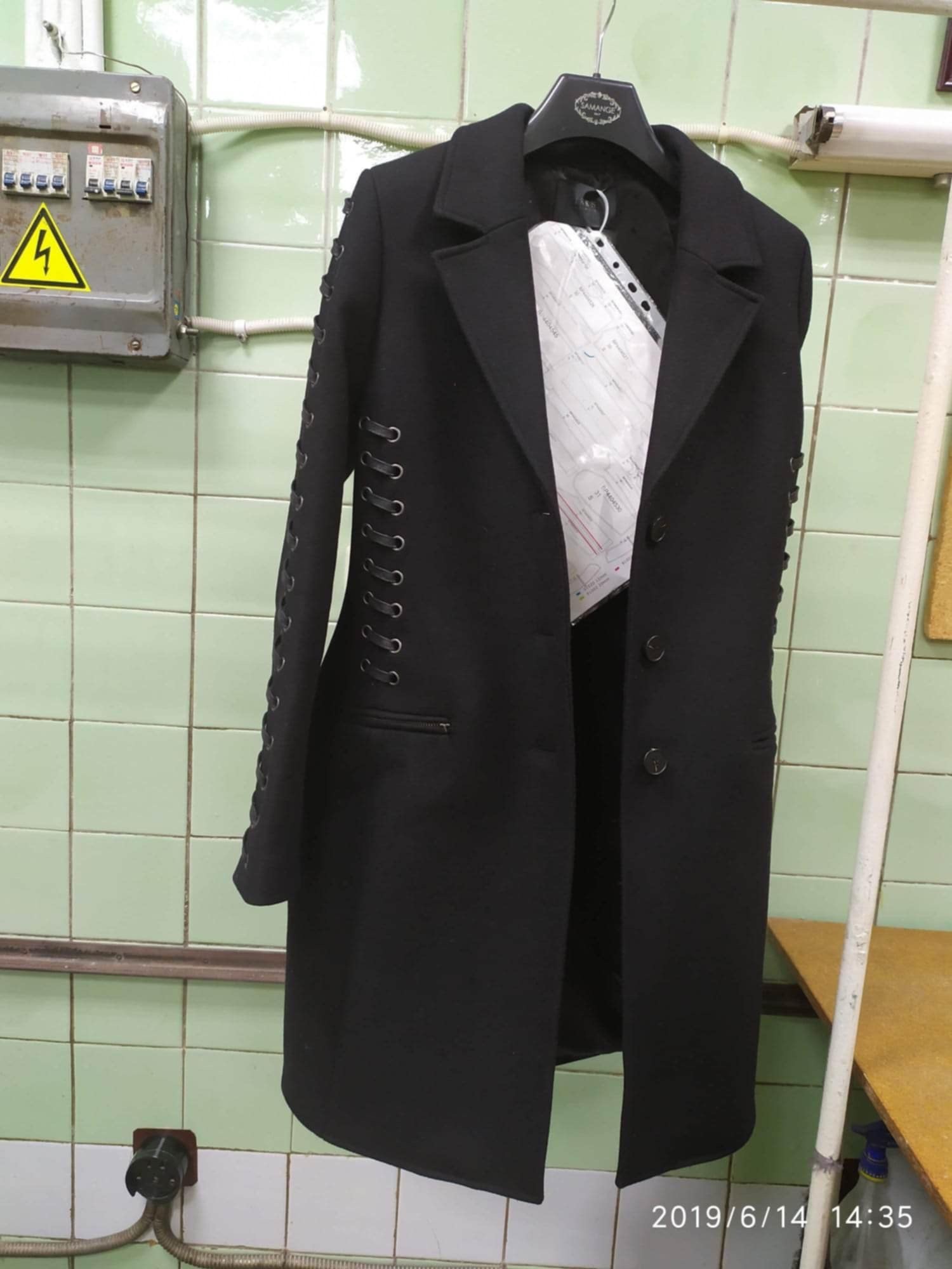 Czarny płaszcz damski z ozdobnym sznurowaniem na rękawach, wiszący na wieszaku w pomieszczeniu z zielonymi kafelkami. Widoczna skrzynka elektryczna i data na dole zdjęcia.