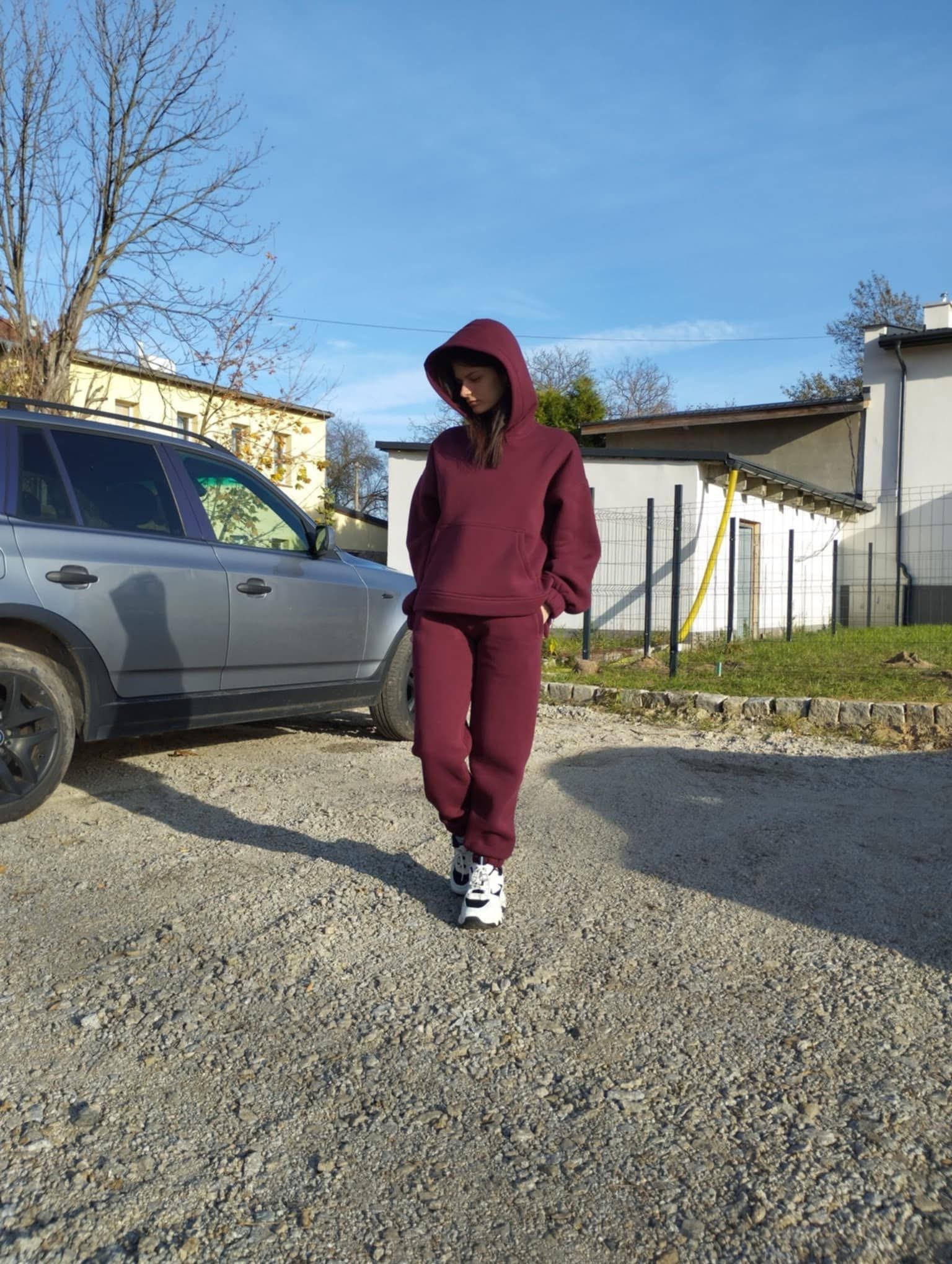 Modelka prezentuje bordowy, dresowy komplet z kapturem na zewnątrz, obok zaparkowanego srebrnego SUV-a, w słoneczny dzień. Widoczne budynki w tle.
