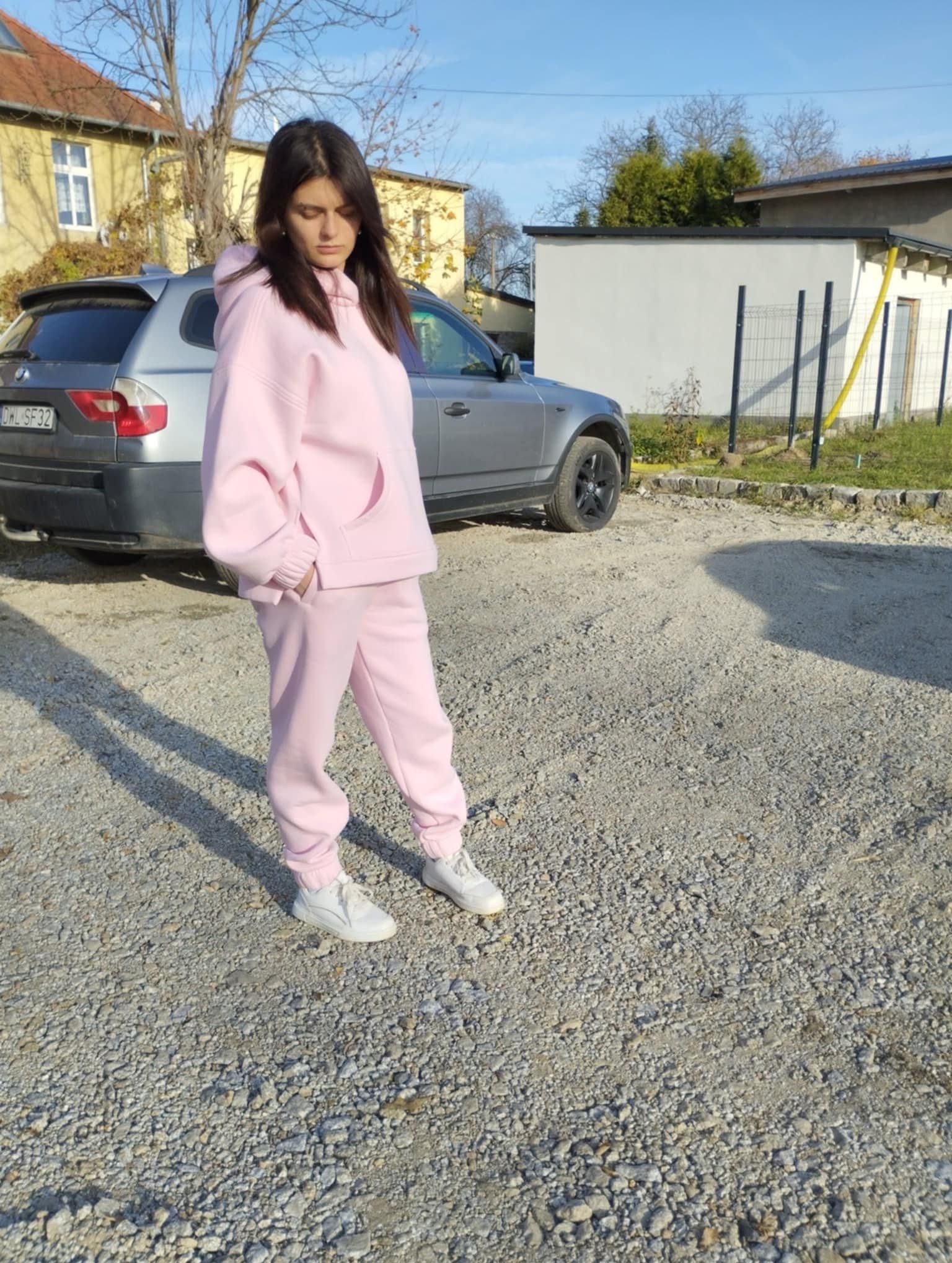 Kobieta w różowym dresie z kapturem, stojąca na żwirowym podłożu, prezentuje komplet odzieży na tle szarego samochodu i zabudowań.