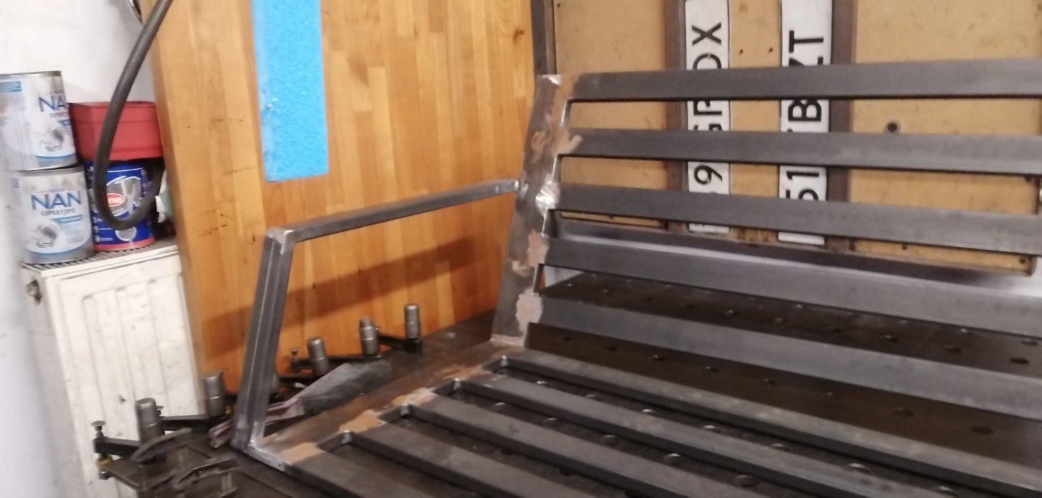 Metalowa konstrukcja w warsztacie na stole spawalniczym, w trakcie tworzenia, z widocznymi śladami spawania i obróbki. W tle blacha i puszki.