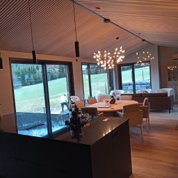 Grzegorz Kaczor - Nowoczesne wnętrze z drewnianym sufitem lamelowym i dużymi oknami, z widokiem na zewnątrz. Wnętrze oświetlone designerskimi lampami wiszącymi.