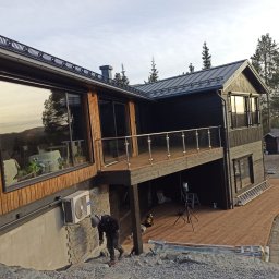 Grzegorz Kaczor - Nowoczesny dom z drewnianym tarasem i szklanymi balustradami. Widoczny pracownik przy wykończeniu terenu. Elementy drewniane w odcieniach brązu i czerni.