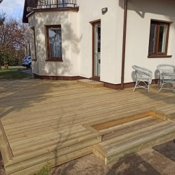 Grzegorz Kaczor - Jasny taras z drewna przed domem z białymi ścianami i brązowymi oknami. Na tarasie wiklinowe krzesła i stolik. Widoczne schody tarasowe i fragment ogrodu.