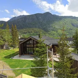 Nowoczesny dom z drewna z dużymi oknami i tarasem, na tle górskiego krajobrazu, częściowo zasłonięty przez rusztowanie.