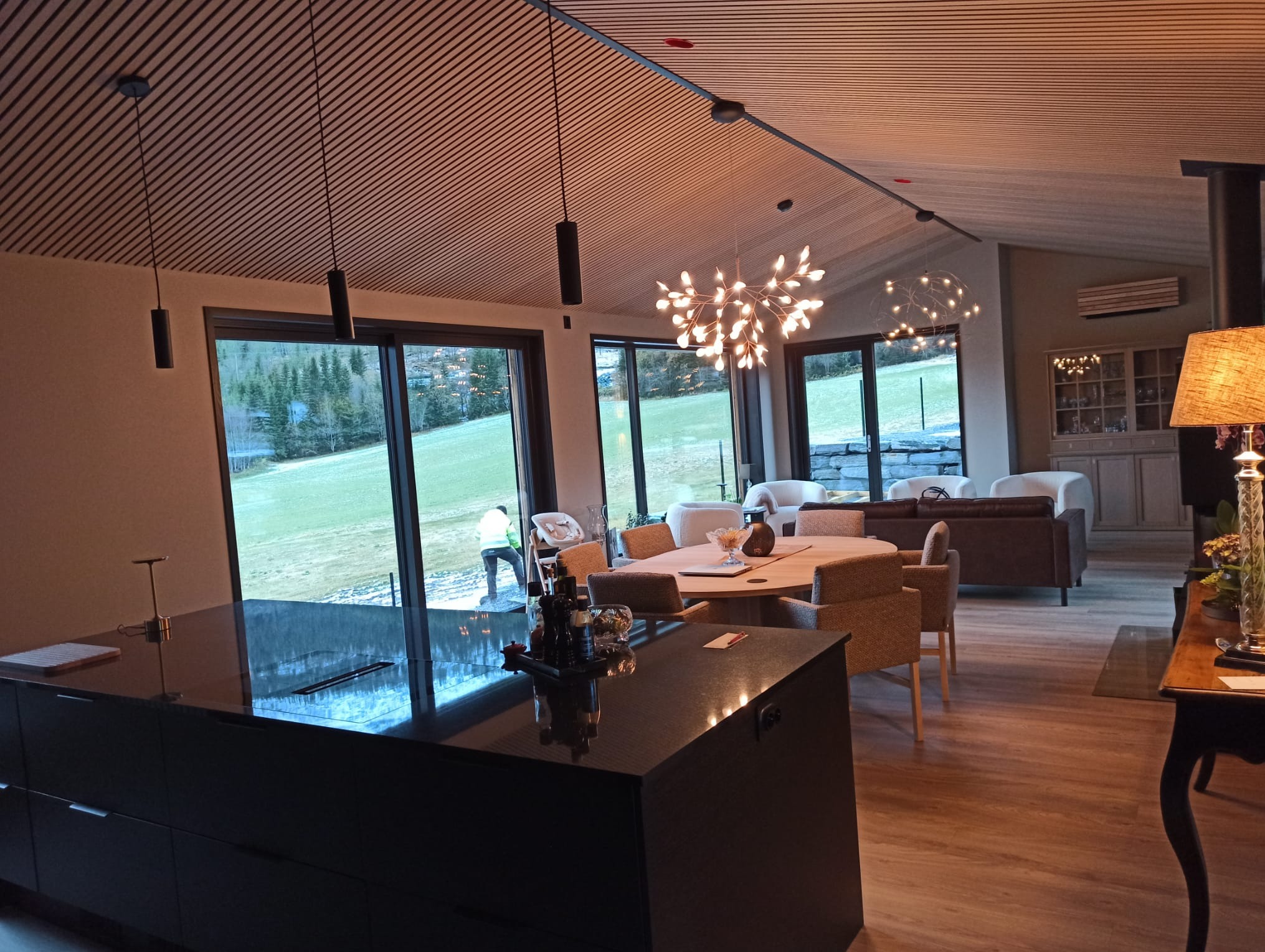 Nowoczesne wnętrze z drewnianym sufitem lamelowym i dużymi oknami, z widokiem na zewnątrz. Wnętrze oświetlone designerskimi lampami wiszącymi.