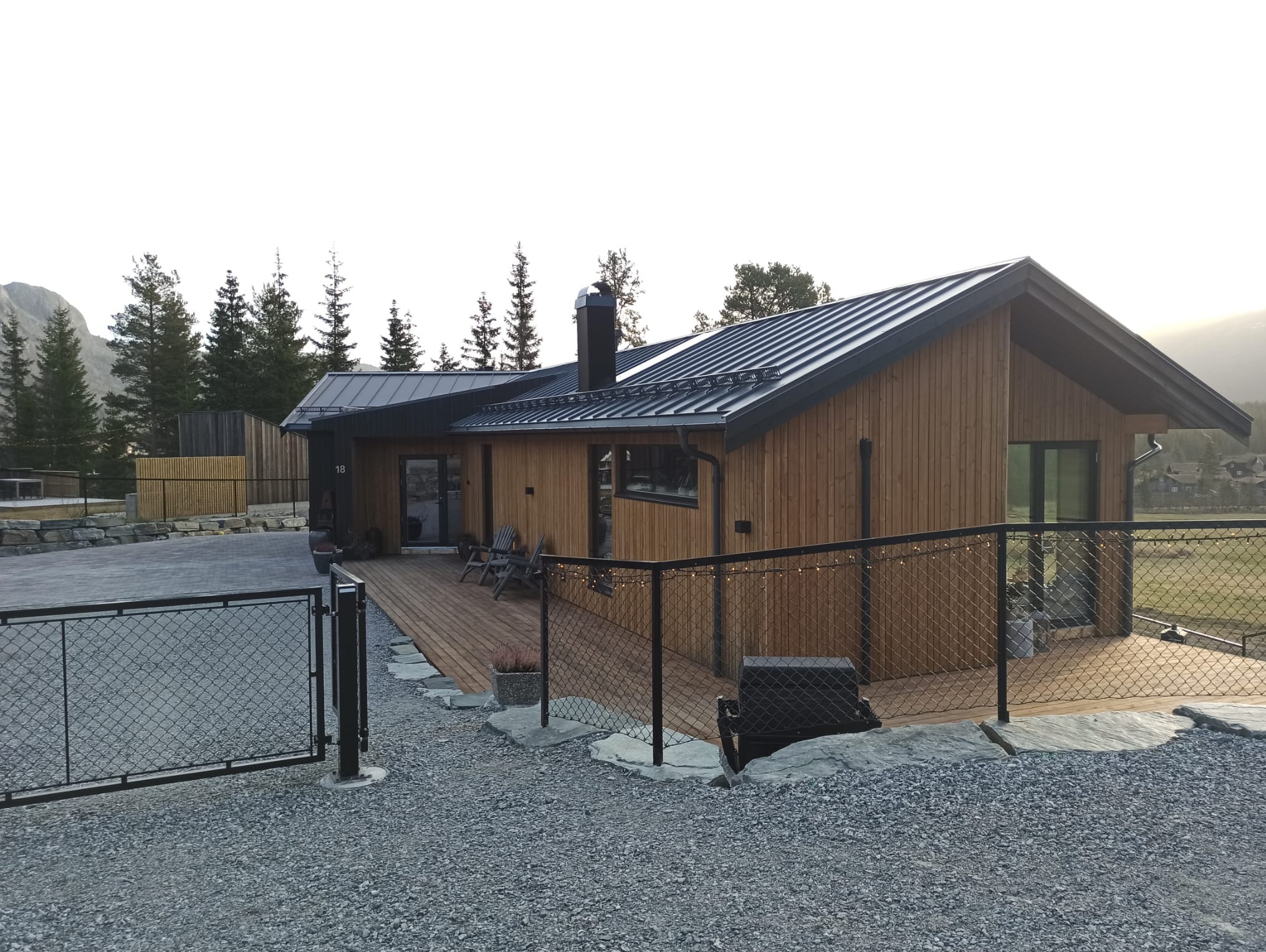 Nowoczesny dom z drewnianą elewacją i tarasem, otoczony żwirowym podłożem i metalowym ogrodzeniem. W tle las iglasty i góry, oświetlone delikatnym, naturalnym światłem.