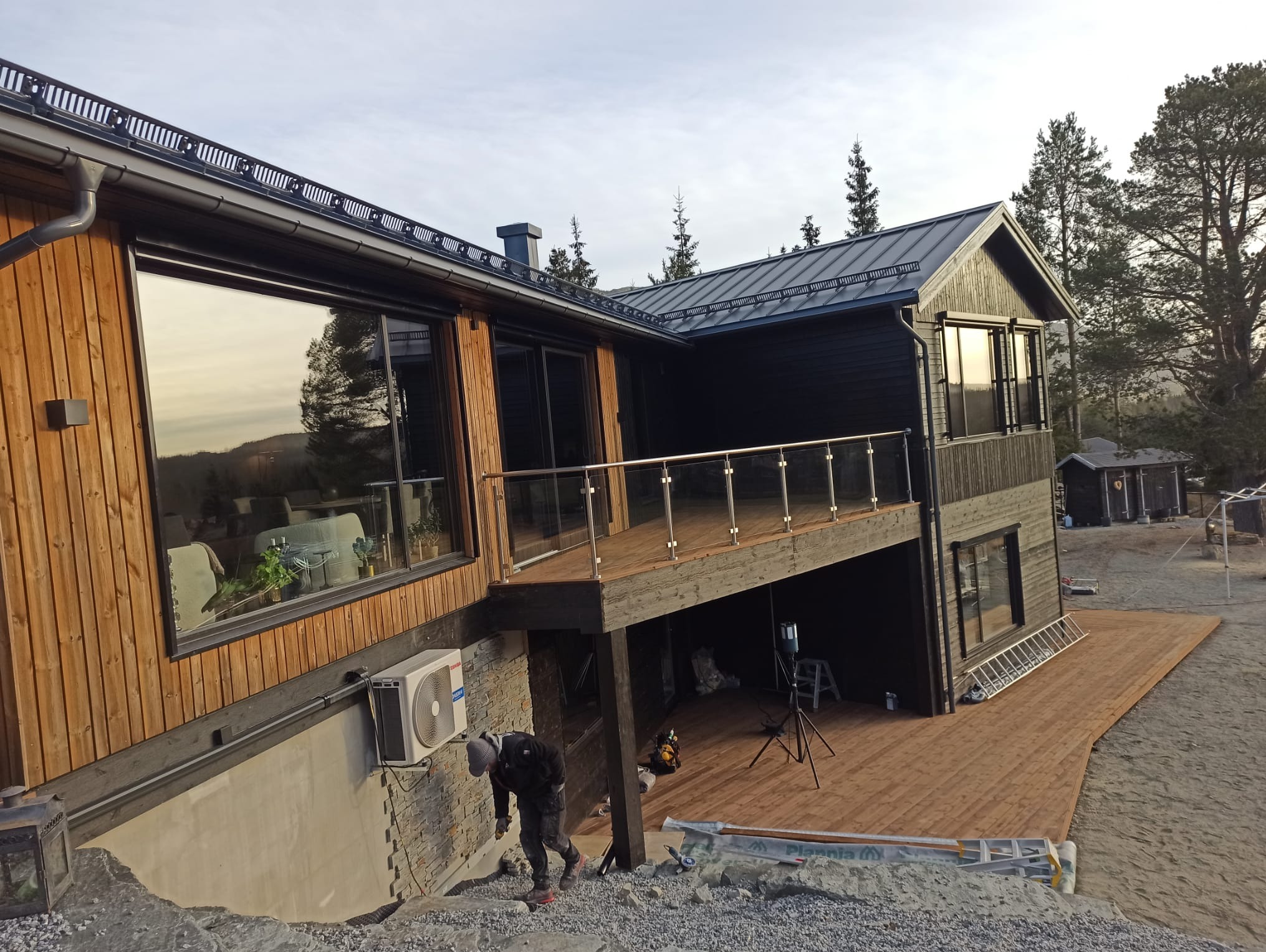 Nowoczesny dom z drewnianym tarasem i szklanymi balustradami. Widoczny pracownik przy wykończeniu terenu. Elementy drewniane w odcieniach brązu i czerni.