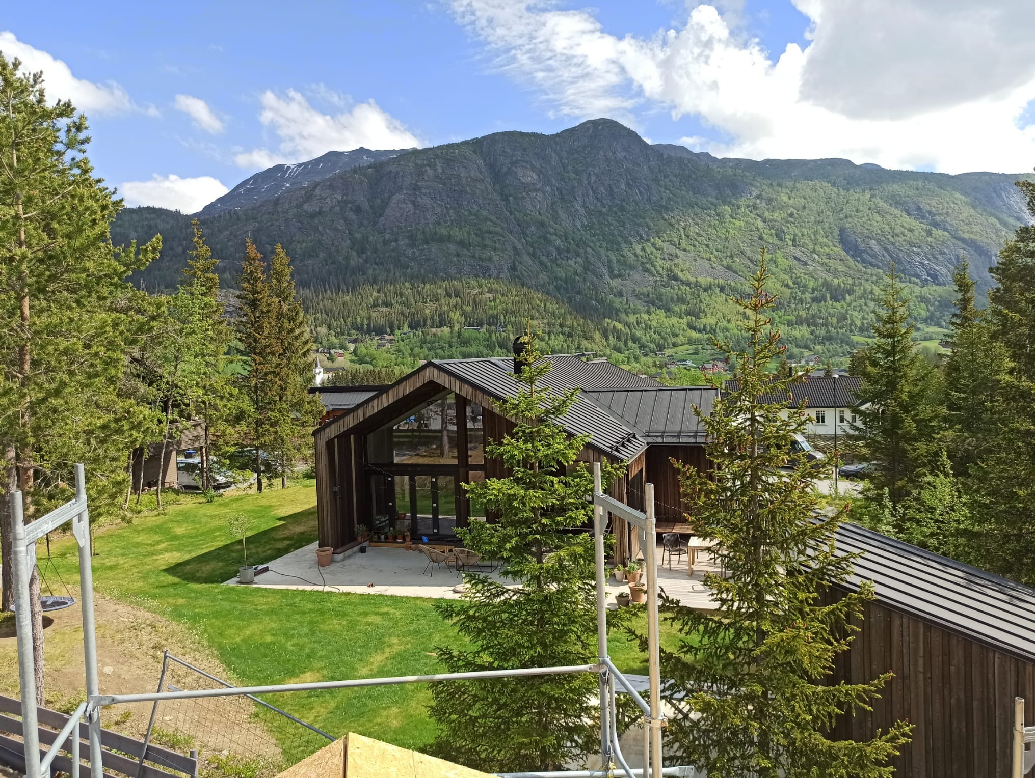 Nowoczesny dom z drewna z dużymi oknami i tarasem, na tle górskiego krajobrazu, częściowo zasłonięty przez rusztowanie.