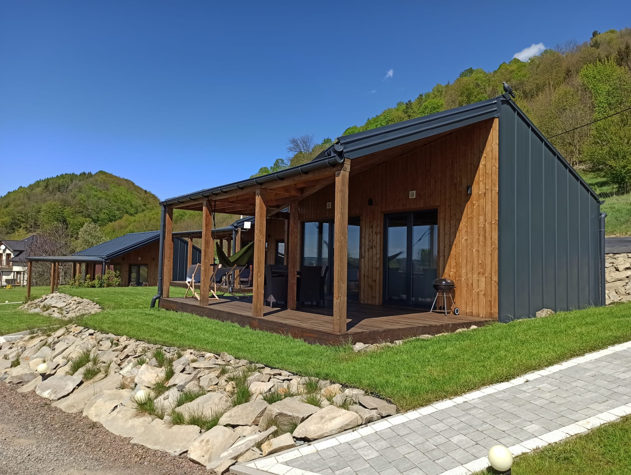 Nowoczesny dom z drewnianą elewacją i tarasem, na którym znajduje się hamak i grill. W tle zielone wzgórza i błękitne niebo.