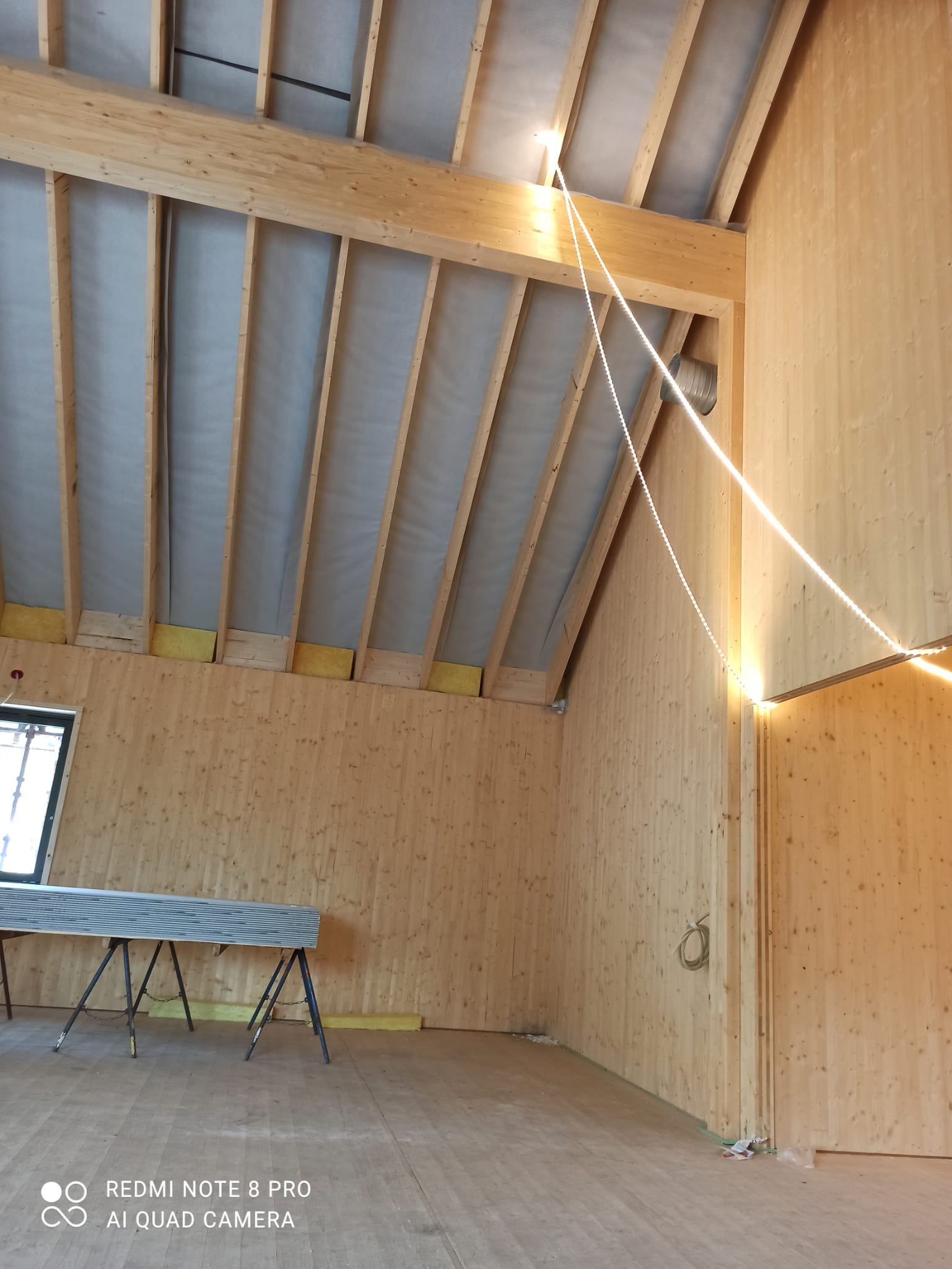 Wnętrze drewnianej konstrukcji w trakcie budowy. Widoczne belki stropowe, ściany z desek, ocieplenie i tymczasowe oświetlenie LED. Na podłodze leżą płyty.