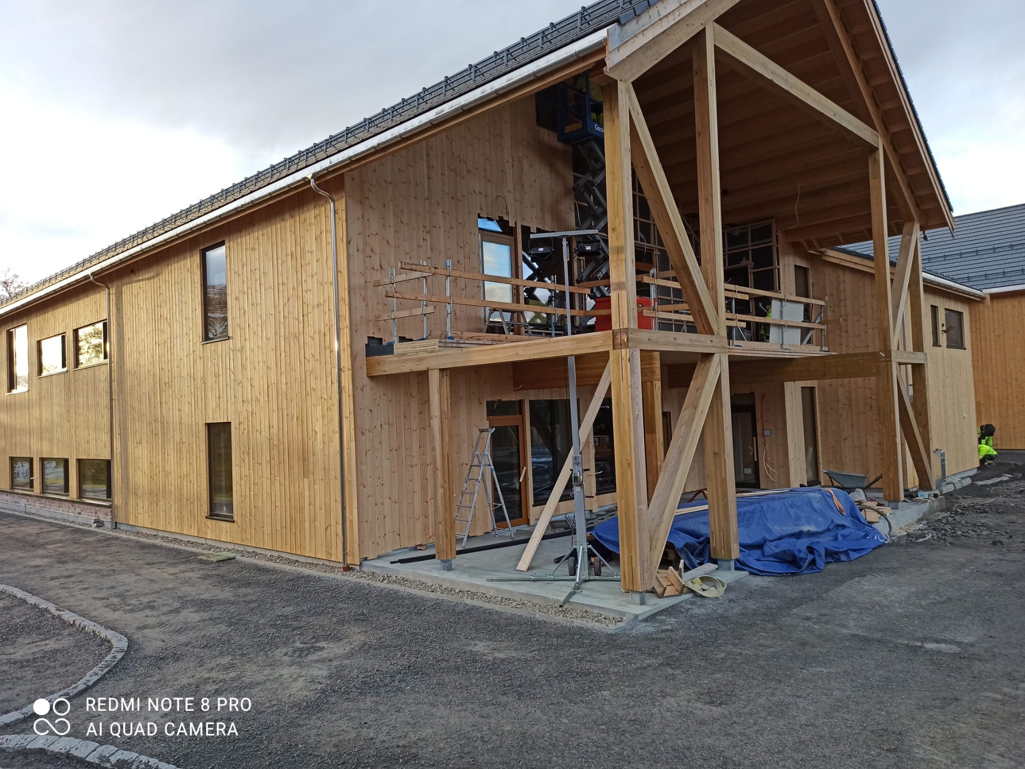 Budowa domu z drewna: surowe ściany, drewniana konstrukcja tarasu na piętrze, drabina i podnośnik w tle. Widok z zewnątrz na plac budowy.