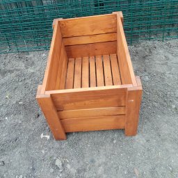 Firma Usługowo Produkcyjna - Drewniana skrzynia na kwiaty z listew, pomalowana na brązowo, stoi na szarej ziemi. W tle zielona siatka ogrodzeniowa. Widok z góry, oświetlenie naturalne.