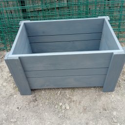 Firma Usługowo Produkcyjna - Szara, drewniana skrzynia na ziemi, z widoczną strukturą drewna i świeżym malowaniem. W tle zielona siatka ogrodzeniowa. Skrzynia wykonana z desek, z solidnymi narożnikami.