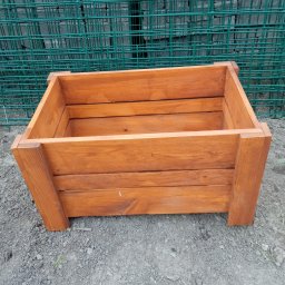 Firma Usługowo Produkcyjna - Lakierowana, drewniana skrzynia na ziemi z zieloną siatką w tle. Widoczne łączenia desek i faktura drewna. Prosty, rustykalny styl.