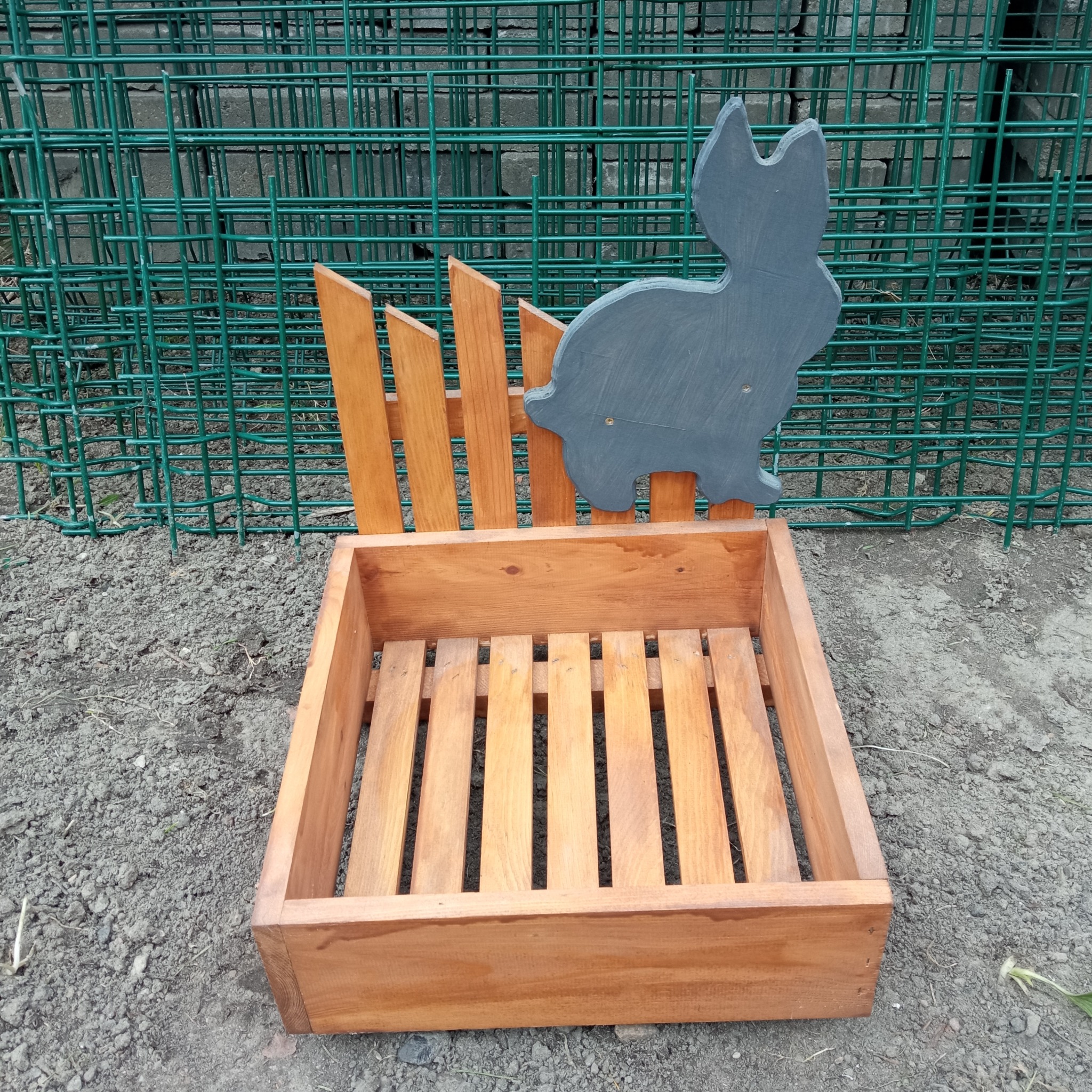 Drewniana skrzynka z ażurowym dnem i dekoracyjnym króliczkiem na tle zielonej siatki. Ręcznie robiona donica ogrodowa z naturalnego drewna.