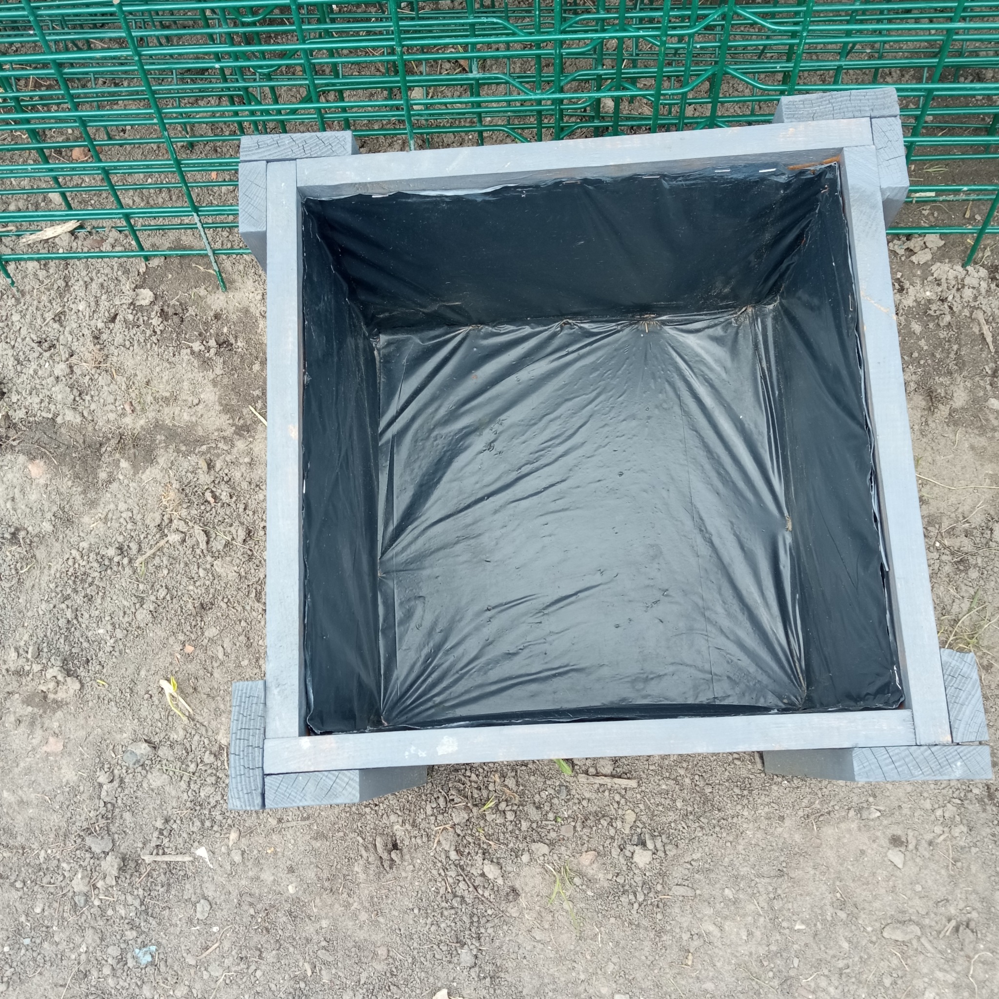 Szara, drewniana skrzynia na ziemi, wyłożona czarną folią, widziana z góry. Elementy konstrukcyjne z widocznym rysunkiem drewna. W tle zielona siatka ogrodzeniowa.