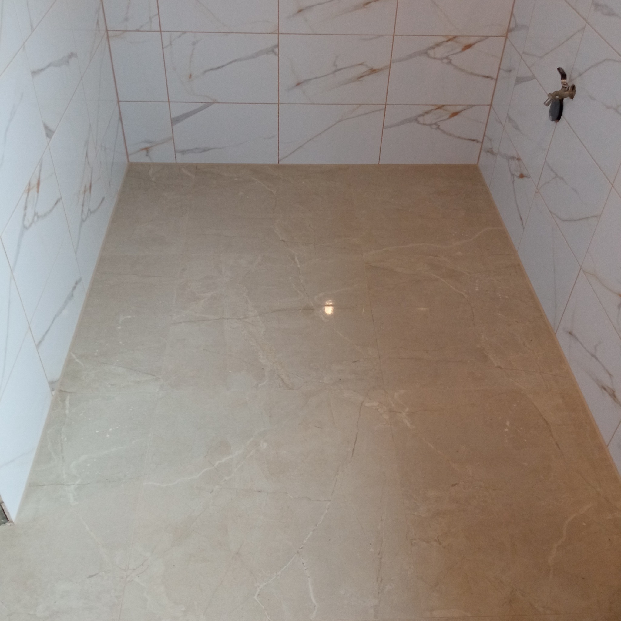 Wykończona łazienka z beżowymi płytkami podłogowymi i białymi płytkami ściennymi z marmurkowym wzorem. Widoczny fragment instalacji wodnej.