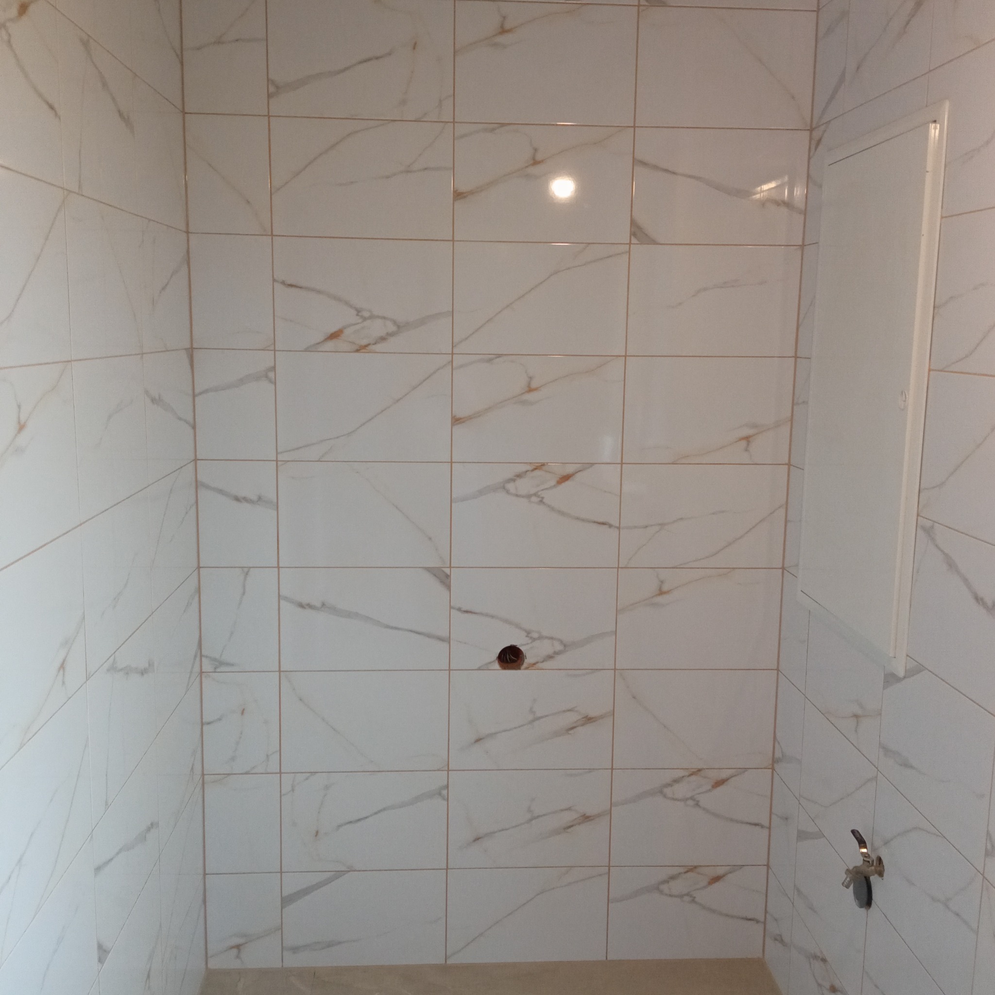 Wykończona łazienka z białymi, kwadratowymi płytkami imitującymi marmur z delikatnym, złotym żyłkowaniem. Widoczny otwór na baterię i zawór.