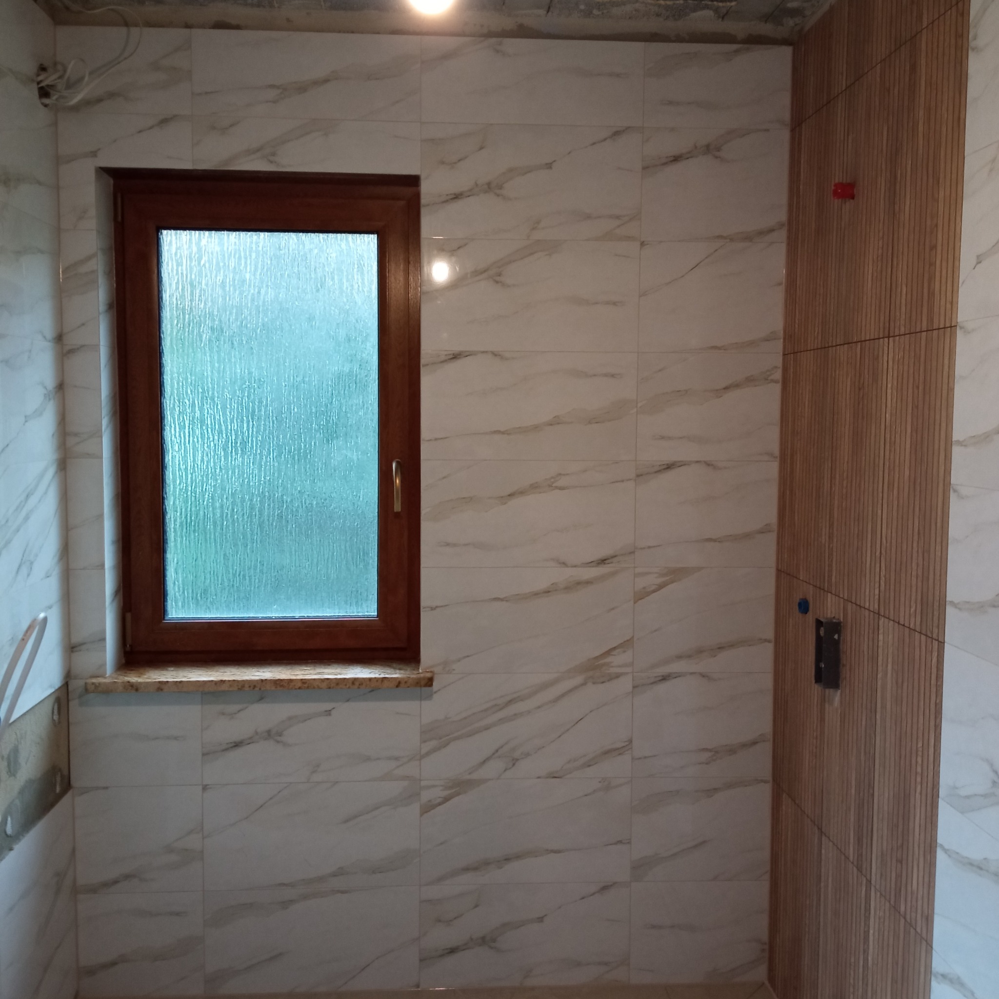 Wykończona łazienka z oknem i panelami ściennymi. Białe płytki imitujące marmur, drewniana rama okienna i pionowe panele z drewna. Widoczne niedociągnięcia wykończeniowe.