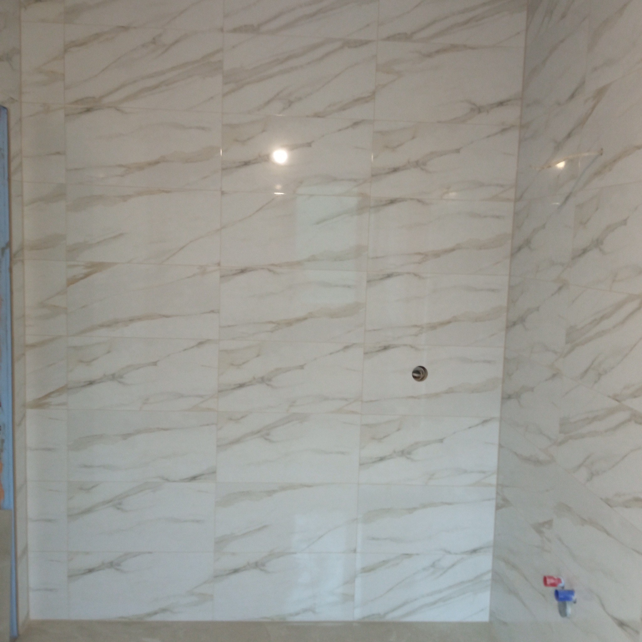 Ściana wyłożona jasnymi, marmurkowymi płytkami ceramicznymi w łazience. Widoczny otwór na armaturę i fragment podłogi. Ujęcie z bliska, prezentujące wzór i fakturę płytek.