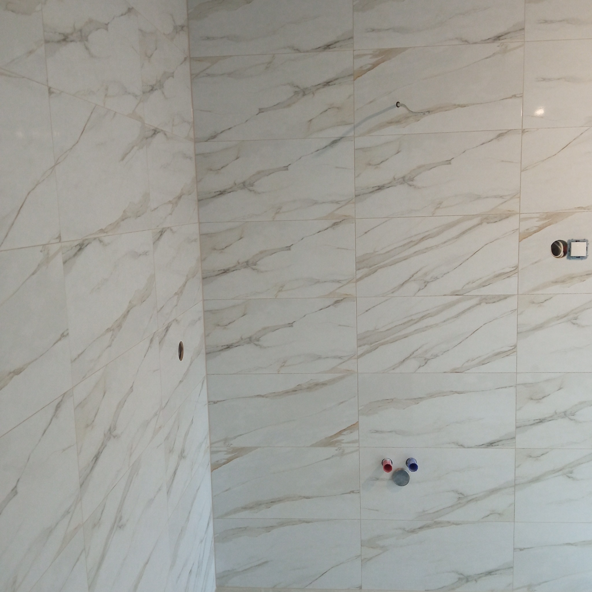 Wykończona ściana łazienki z białych, marmurkowych płytek. Widoczne otwory na armaturę i gniazdko. Ujęcie z bliska, prezentujące detale wzoru i fug.