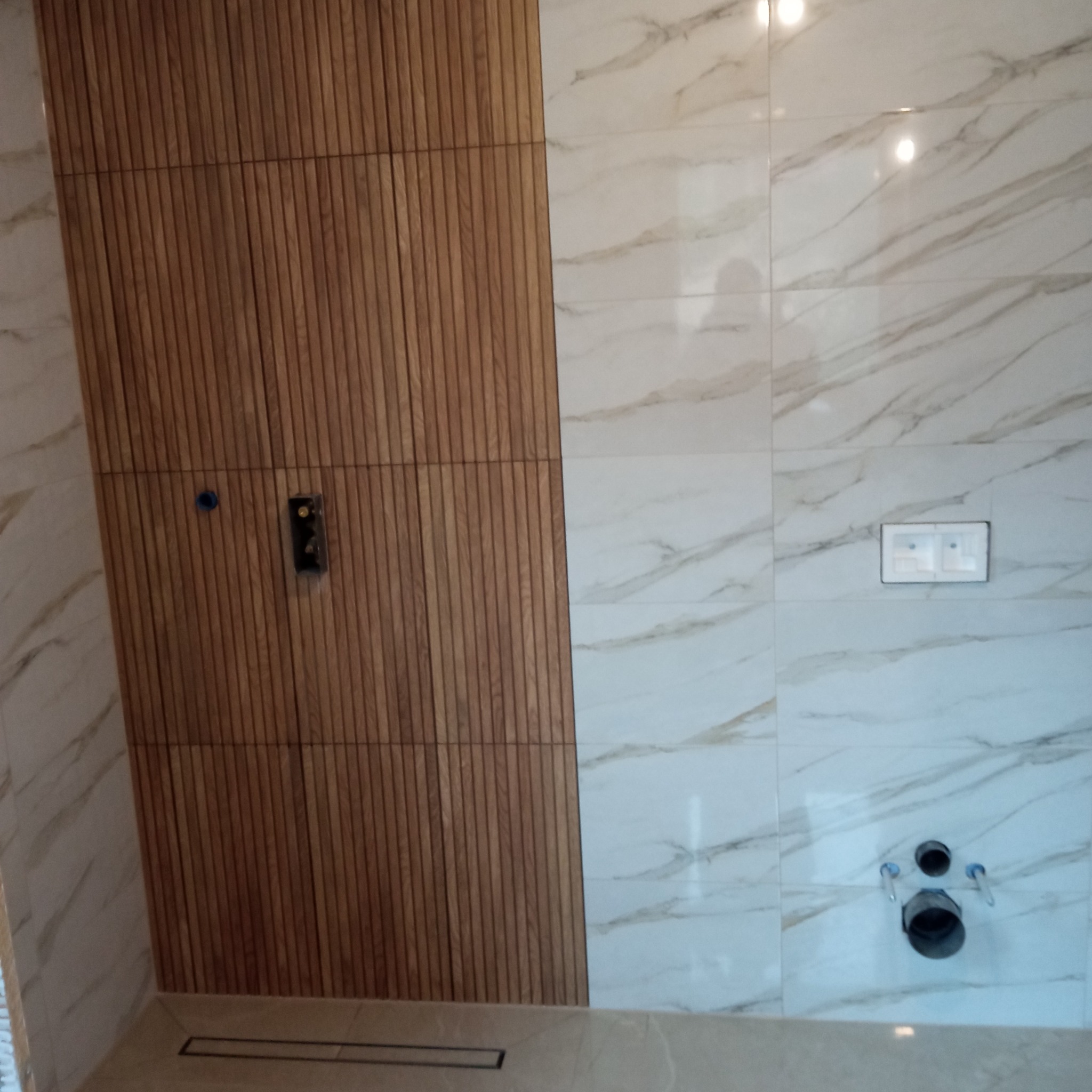 Wykończona ściana w łazience z drewnianymi panelami i marmurowymi płytkami. Widoczne elementy instalacji sanitarnej i elektrycznej. Nowoczesny design.