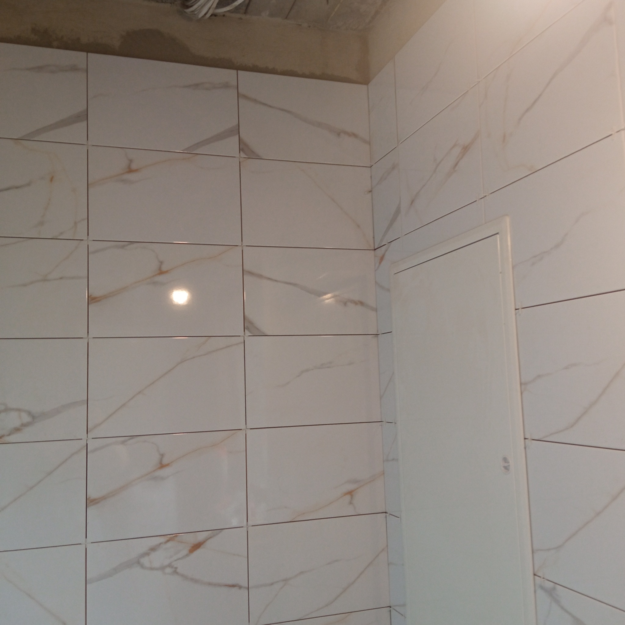 Wykończona ściana w łazience z białymi płytkami imitującymi marmur, z widoczną białą klapą rewizyjną. Widoczne krzyżyki dystansowe w spoinach.