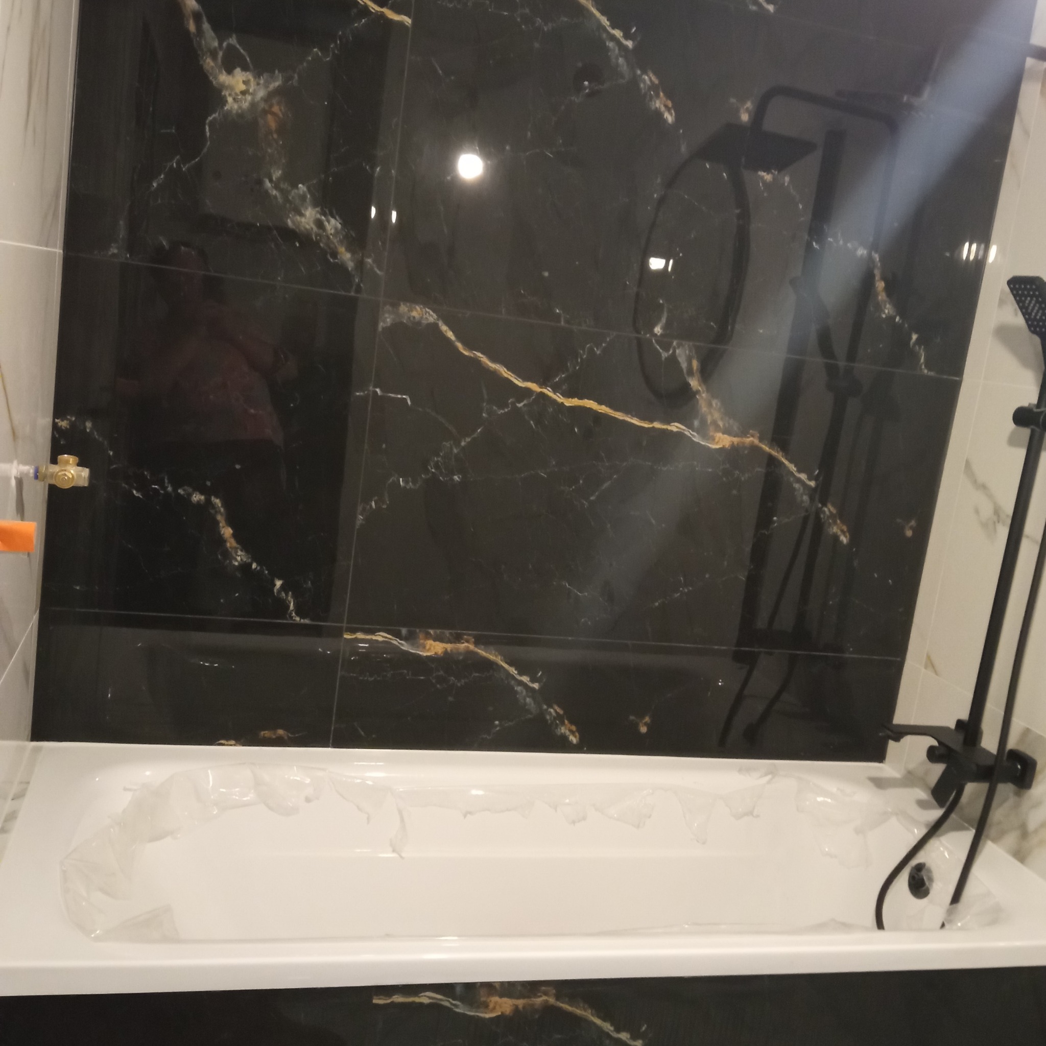 Wykończona łazienka z czarnymi płytkami imitującymi marmur i białą wanną zabezpieczoną folią. Nowoczesna armatura w kolorze czarnym.