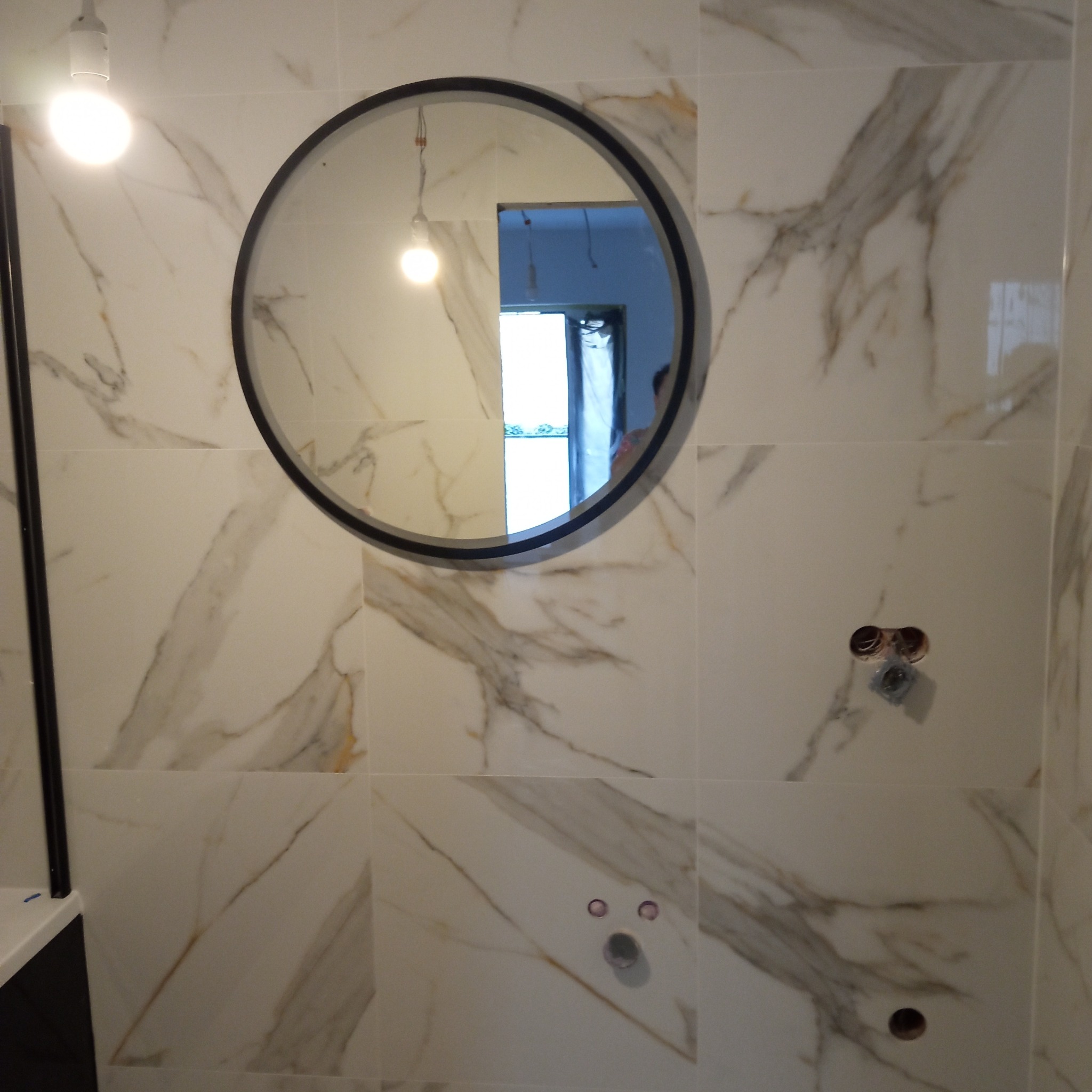 Wykończona ściana łazienki z białych płytek imitujących marmur, z okrągłym lustrem w czarnej ramie. Widoczne gniazdka elektryczne i oświetlenie.