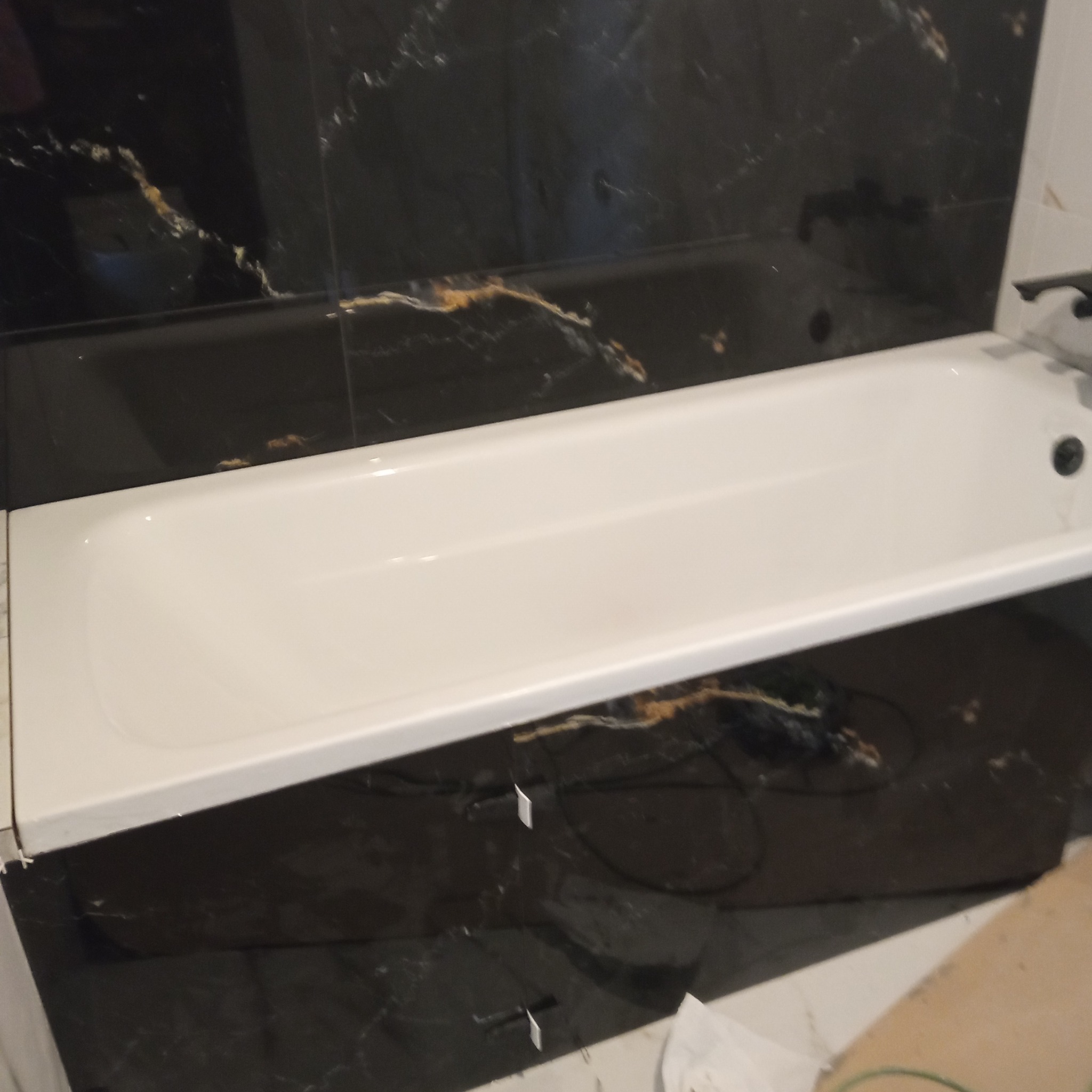 Biała wanna w łazience z czarnymi płytkami imitującymi marmur. Widoczne fugi i fragment podłogi. Ujęcie z góry, podkreślające kontrast kolorów i faktur.