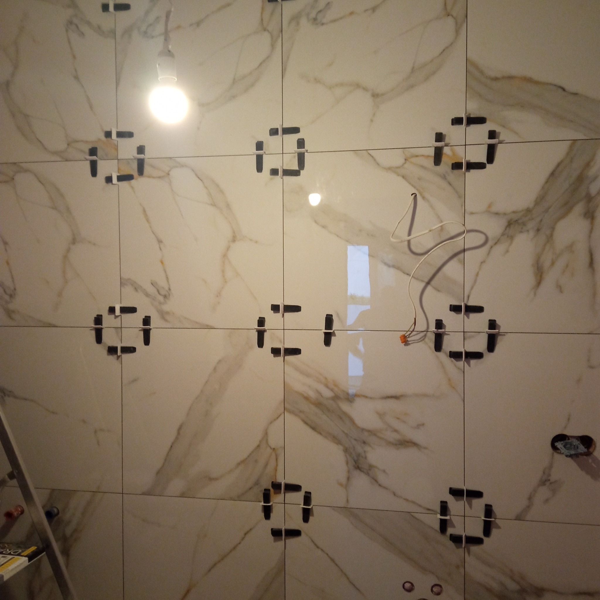 Układanie płytek imitujących marmur na ścianie z użyciem klipsów poziomujących. Widoczne przewody elektryczne i drabina. Oświetlenie halogenowe.
