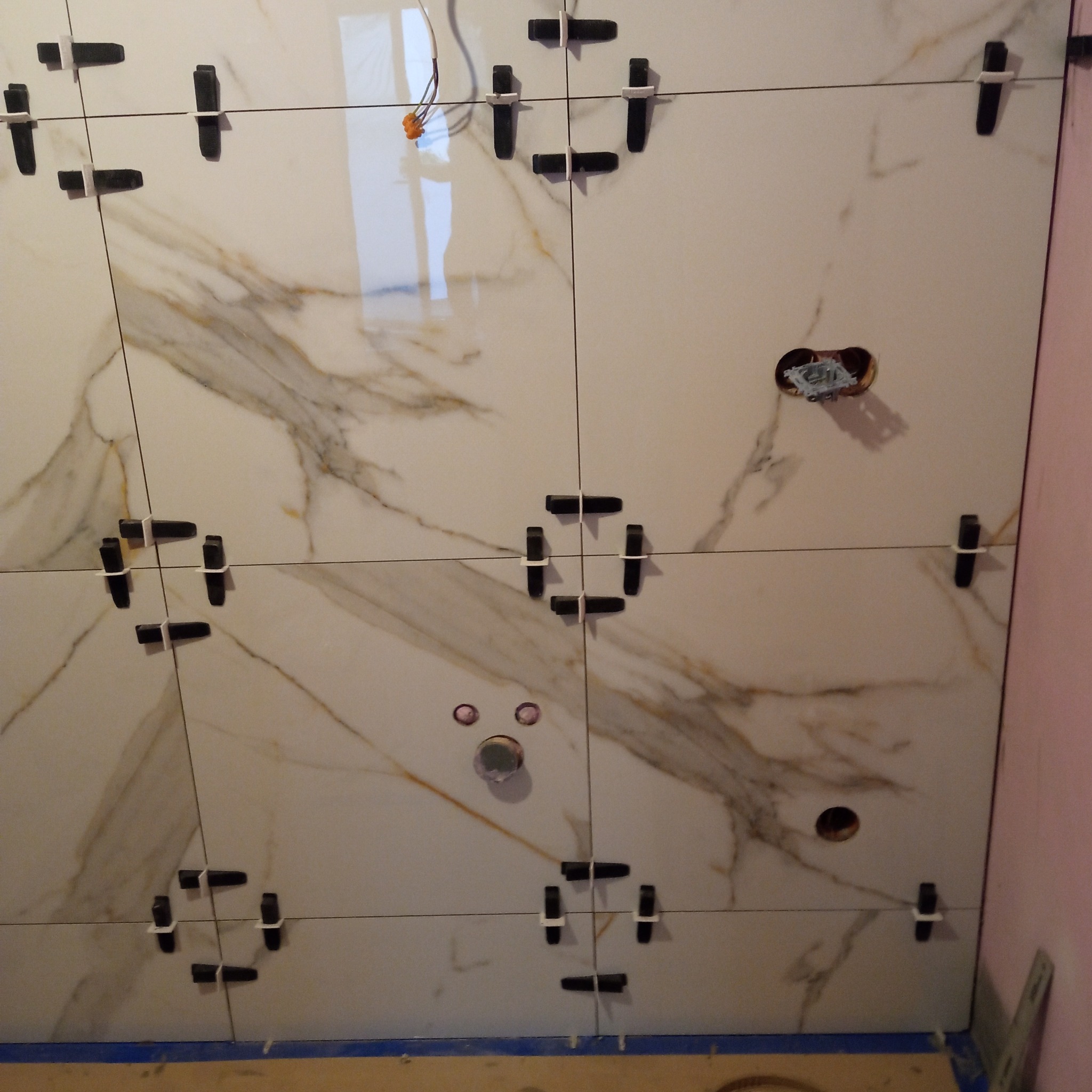 Układanie płytek imitujących marmur na ścianie z użyciem klipsów poziomujących. Widoczne otwory na instalacje i fragment różowej ściany.