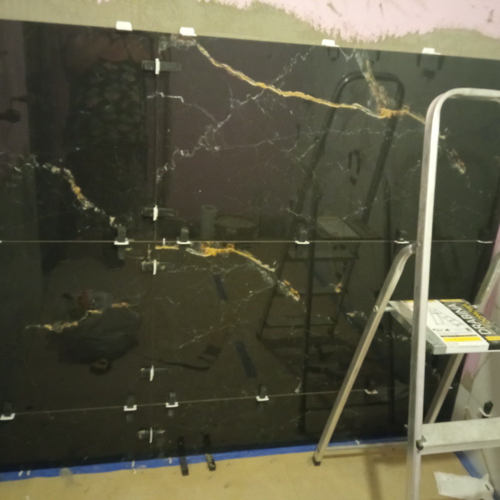 Układanie czarnych, marmurkowych płytek z użyciem krzyżyków dystansowych. Widoczna drabina i fragment ściany. Praca w toku, etap montażu płytek.