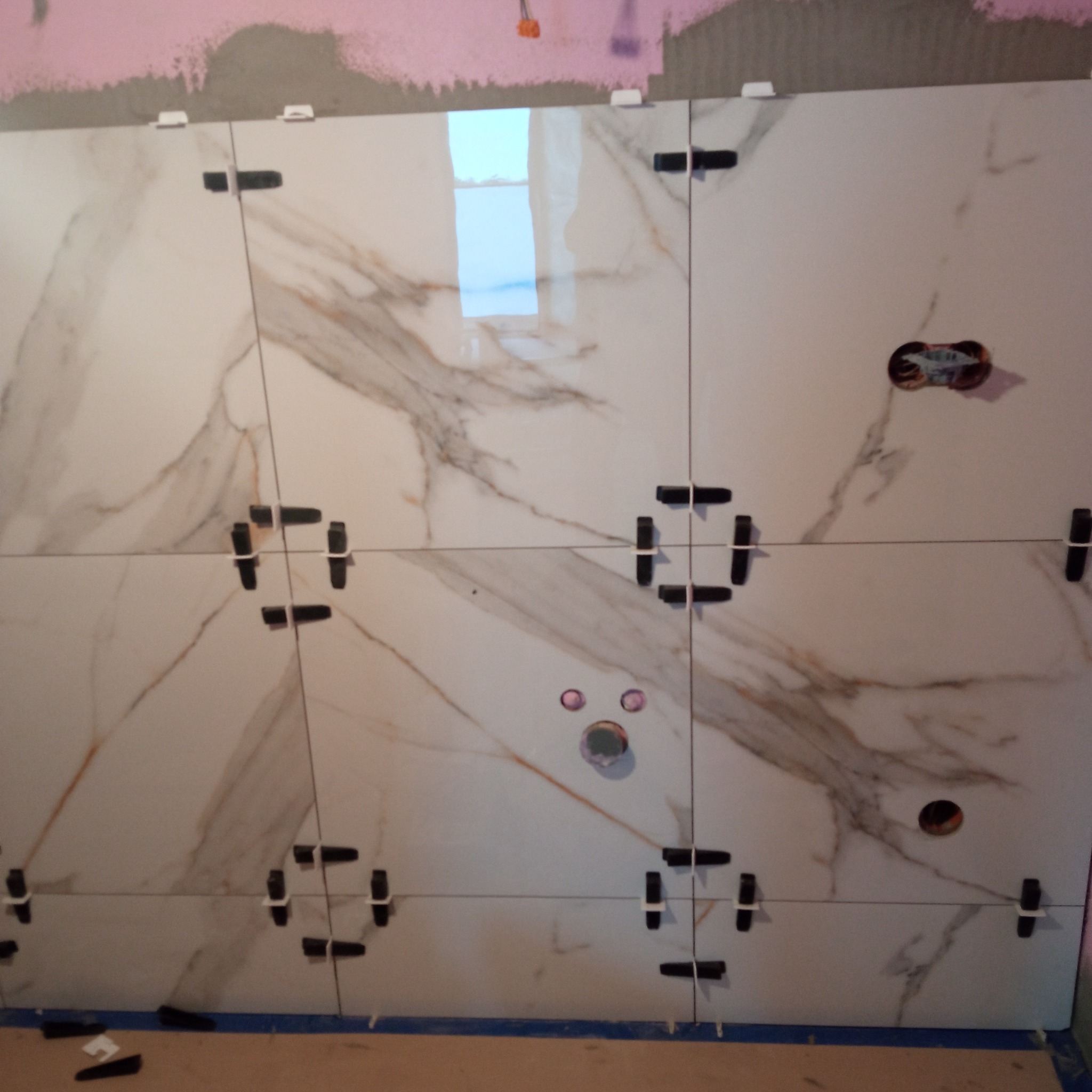 Ściana z płytek imitujących marmur, z czarnymi krzyżykami dystansowymi i otworami na instalacje, w trakcie montażu. Widoczne fragmenty różowej ściany i niebieskiej taśmy.