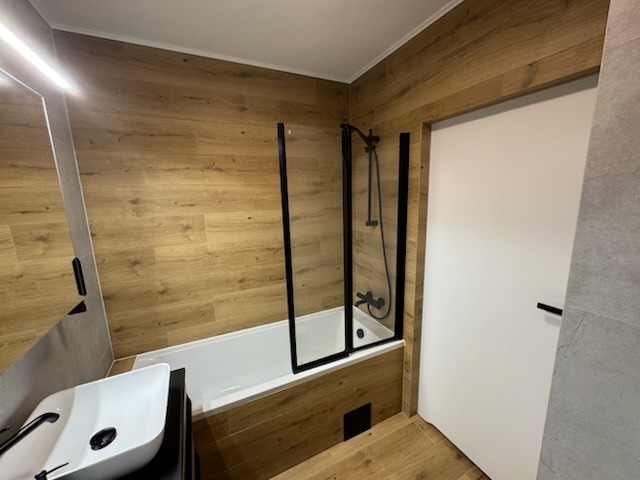 Nowoczesna łazienka z wanną za czarną szybą, umywalką nablatową i drewnopodobnymi płytkami na ścianach. Minimalistyczny design, stonowana kolorystyka.