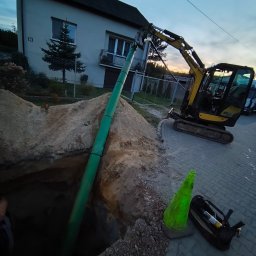 ŻUK Maciej Żukowski - Żółta koparka Yanmar umieszcza zieloną rurę w wykopie na tle domu jednorodzinnego. Widoczny stożek ostrzegawczy i torba narzędziowa.