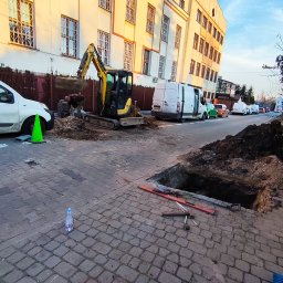 ŻUK Maciej Żukowski - Żółta mini koparka przy wykopie na ulicy z kostki, w tle budynek i zaparkowane samochody. Widoczne narzędzia i akcesoria budowlane.