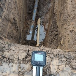 ŻUK Maciej Żukowski - Wykop z rurami i wodą, widoczny detektor podziemnych instalacji. Ujęcie z góry, z butami osoby wykonującej pomiar. Błoto na ziemi i urządzeniu.