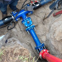 ŻUK Maciej Żukowski - Niebieski trójnik z rurami w wykopie, połączony z czerwoną rurą. Widoczne narzędzia i fragment drabiny. Instalacja wodociągowa w piasku.